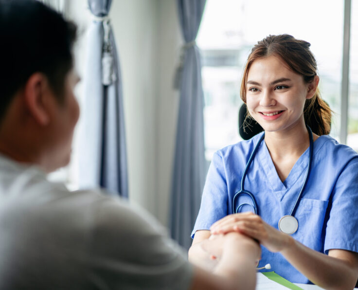 A woman in a blue scrubs is talking to a man. Scene is friendly and caring